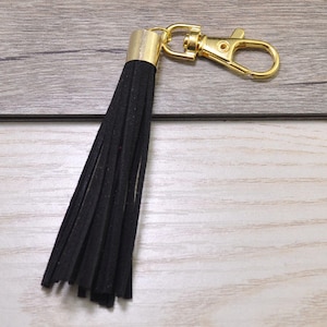 Puede incluir: Un llavero de borla negro con un anillo de llave de metal dorado. La borla está hecha de finos hilos negros de tela.