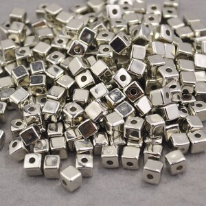 Puede incluir: Un montón de cuentas cúbicas plateadas y brillantes. Cada cuenta tiene un agujero perforado en el centro, y los cubos están dispersos sobre una superficie neutra. Estas cuentas son adecuadas para la fabricación de joyas y manualidades.