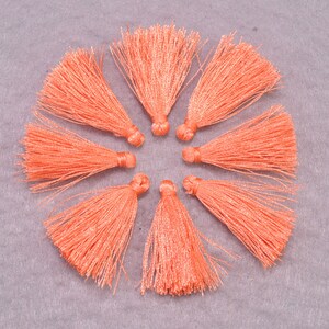 May include: Eight peach-coloured fabric tassels arranged in a circle on a grey background. The tassels are approximately 5 cm long.