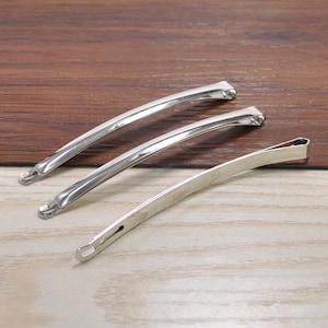 Puede incluir: Tres pinzas para el pelo de metal plateado. Las pinzas son todas del mismo tamaño y forma, y están dispuestas en fila sobre una superficie de madera.