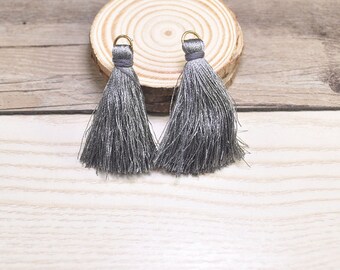 May include: Two gray tassel trim with gold metal rings. The tassels are on a wooden surface.