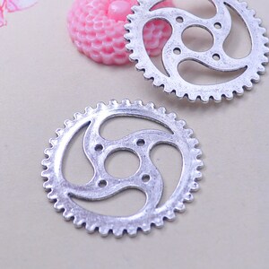 Op de afbeelding: Twee zilveren metalen tandwielen met een decoratief, draaiend ontwerp. De tandwielen hebben een verweerde, antieke afwerking.