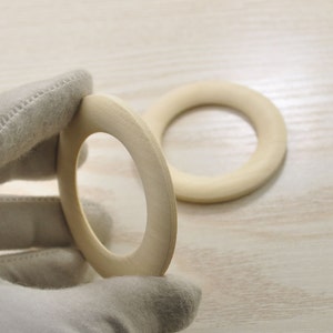 May include: Two unfinished wooden rings, one held in a gloved hand, the other lying on a light wood surface.