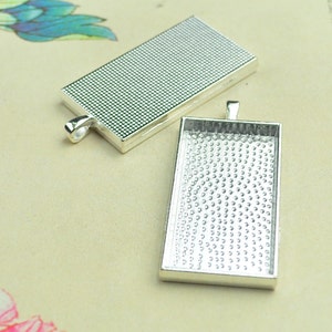 May include: Two silver-toned rectangular pendant trays with a textured surface. The trays are designed to hold a small image or other decorative element.