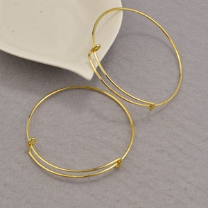 May include: Two gold-toned metal bangle bracelets with a spring-loaded clasp. The bracelets are on a gray surface.