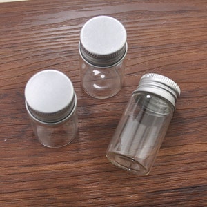 May include: Three small, clear glass vials with silver metal screw-on lids. The vials are cylindrical and empty, suitable for storing small items. The lids are flat and have a ridged edge for easy gripping.