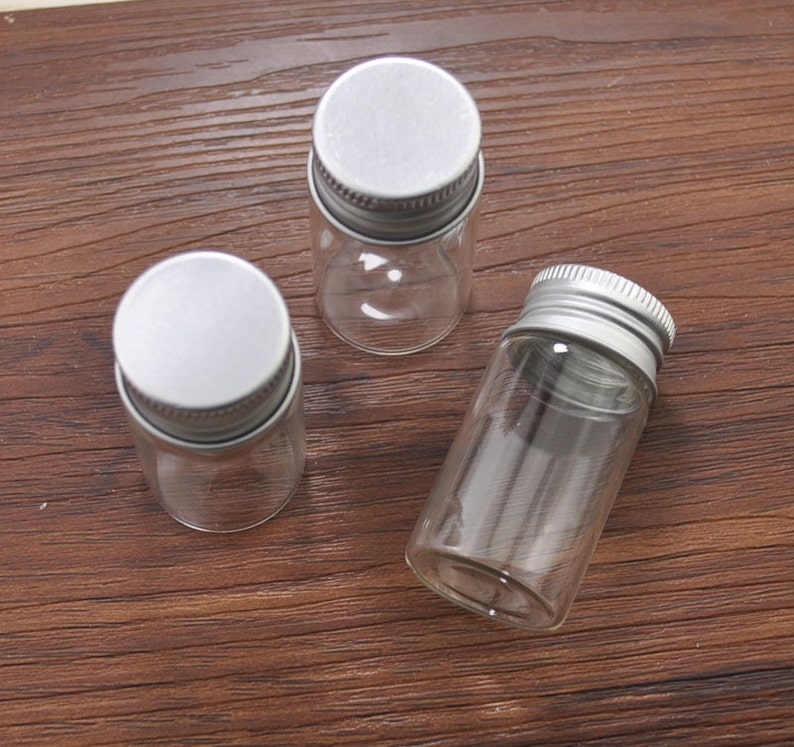 May include: Three small, clear glass vials with silver metal screw-on lids. The vials are cylindrical and empty, suitable for storing small items. The lids are flat and have a ridged edge for easy gripping.