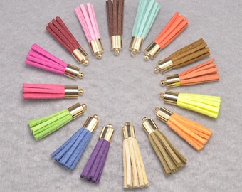 Puede incluir: Surtido de 15 borlas de gamuza de colores vivos con tapas de metal dorado. Las borlas están dispuestas en círculo sobre una superficie gris.