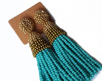 May include: A pair of turquoise beaded tassel earrings with gold accents. The earrings are hanging from a brown card.