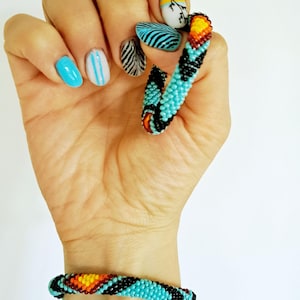 May include: A hand shows a beaded bracelet and a broken beaded ring, both in turquoise, black, orange, and yellow. The nails are painted with various designs, including blue, white, and black patterns.