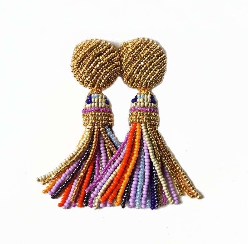 Puede incluir: Un par de pendientes de borla de cuentas doradas con cuentas multicolores en tonos de morado, naranja, azul y crema.