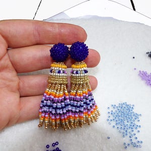 Puede incluir: Un par de pendientes de borla de cuentas con un esquema de color morado y dorado. Los pendientes tienen una parte superior redonda de cuentas y una borla larga y con flecos.