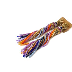 Puede incluir: Un par de pendientes largos de cuentas de colores con tapas doradas. Los pendientes est&aacute;n hechos con una variedad de colores, incluyendo morado, azul, naranja y dorado.