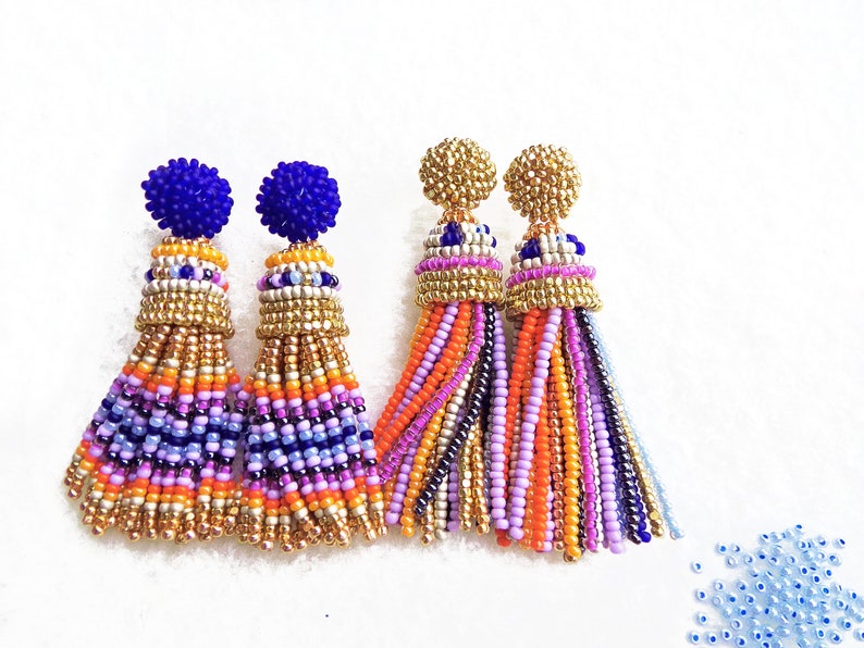 Puede incluir: Un par de pendientes de borla de cuentas de colores con detalles dorados. Las borlas est&aacute;n hechas de peque&ntilde;as cuentas en tonos de morado, rosa, naranja y azul. La parte superior de los pendientes es azul y dorada.