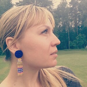 Puede incluir: Una mujer con cabello rubio que lleva una camisa gris oscura y pendientes de borla azul y dorado. Los pendientes tienen una cuenta redonda azul en la parte superior y una borla larga con cuentas doradas, blancas y rosadas.