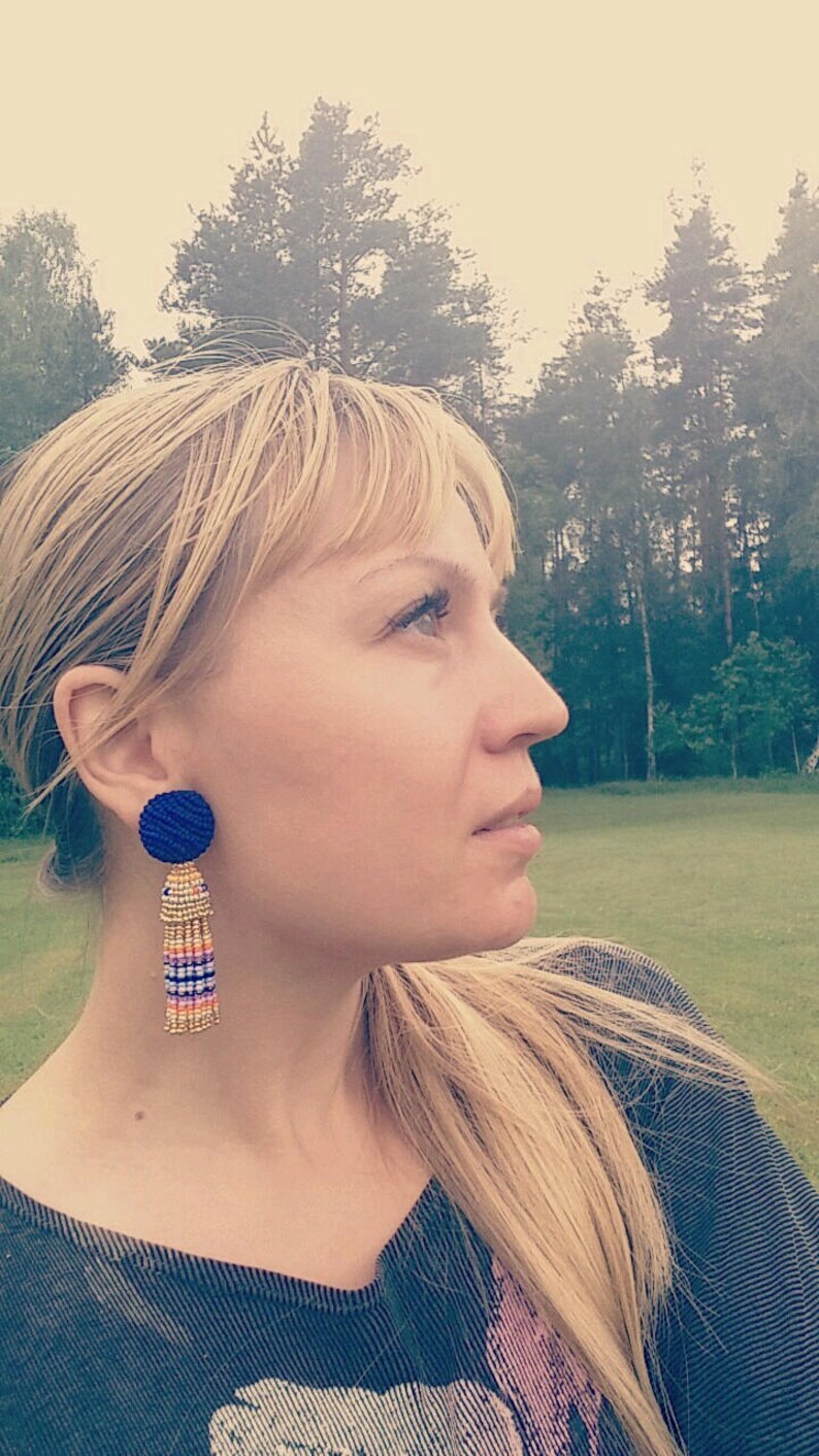 Puede incluir: Una mujer con cabello rubio que lleva una camisa gris oscura y pendientes de borla azul y dorado. Los pendientes tienen una cuenta redonda azul en la parte superior y una borla larga con cuentas doradas, blancas y rosadas.