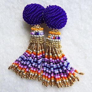 Puede incluir: Un par de pendientes de cuentas moradas con borlas de colores. Las borlas est&aacute;n hechas de cuentas doradas, naranjas, rosas y moradas.