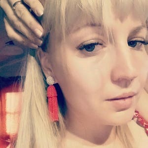 May include: A woman with blonde hair wearing red tassel earrings. The earrings are made of small beads and have a silver charm at the top.