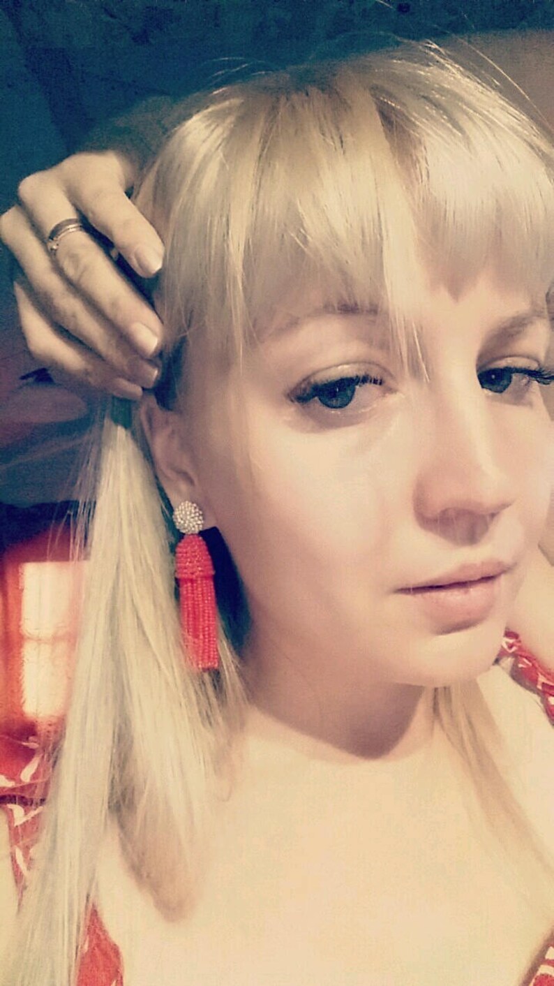 May include: A woman with blonde hair wearing red tassel earrings. The earrings are made of small beads and have a silver charm at the top.
