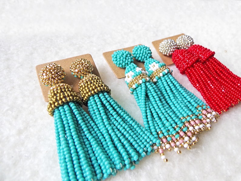 May include: A pair of turquoise beaded tassel earrings with gold accents. The earrings are shown with two other pairs of beaded tassel earrings in red and white.