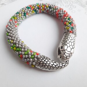 May include: A silver snake bracelet with a beaded body in shades of gray, green, yellow, and orange. The snake's head is a silver charm with a textured surface.