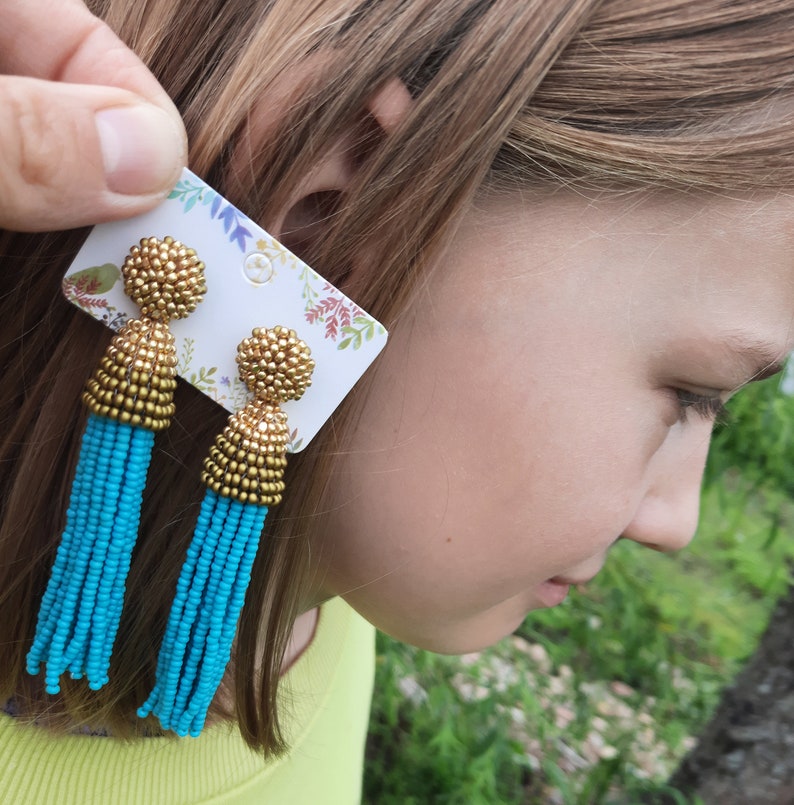 May include: A pair of turquoise tassel earrings with gold accents. The earrings are on a white card with floral designs.