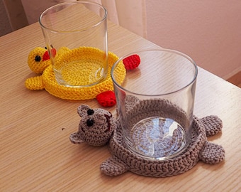May include: Two crocheted coasters, one shaped like a duck and the other like a bear, with glasses on top. The duck coaster is yellow with red accents, and the bear coaster is gray with black eyes.