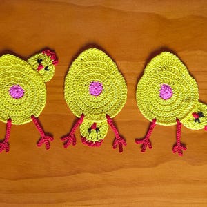 May include: Three crocheted yellow chick coasters with pink centers and red legs. The chicks are arranged in a row on a wooden surface.