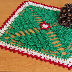 May include: A green crocheted Christmas tree doily with a white and red border. The doily is on a wooden surface with a pine cone next to it.