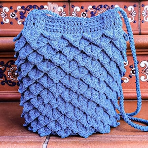 May include: A blue crocheted bag with a scale pattern and a long strap. The bag is sitting on a tiled surface.