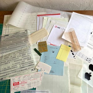 May include: A collection of vintage papers and ephemera, including graph paper, printed sheets, and a clear plastic ruler. There are also several small cards, a circuit board, and two Apple logo stickers. The items are arranged on a wooden surface.