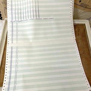 May include: Vintage computer printout paper with horizontal green and white stripes and small blue numbers and symbols. The paper has perforated edges and is displayed on a white surface.