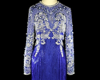 May include: A long-sleeved, royal blue gown with a fitted bodice and a flowing skirt. The gown is covered in intricate beading and sequins, creating a shimmering effect. The bodice features a high neckline and a delicate pattern of floral motifs.