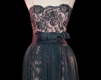 Puede incluir: Un vestido de encaje negro con escote de corazón y un lazo de satén negro en la cintura. El vestido está en un maniquí blanco sobre un fondo negro.