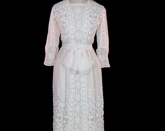 May include: A white, floor-length dress with intricate lace detailing and a bow at the waist.