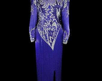 May include: A long, royal blue gown with intricate silver sequin and bead embellishments. The dress features a high neckline, long sleeves, and a dramatic split in the skirt. The bodice has a sheer panel with silver detailing. The dress is displayed on a mannequin.