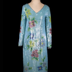 Könnte beinhalten: Ein langärmeliges, hellblaues Paillettenkleid mit einem Blumenmuster in den Farben Rosa, Gelb, Grün und Blau. Das Kleid hat einen V-Ausschnitt und eine figurbetonte Silhouette.
