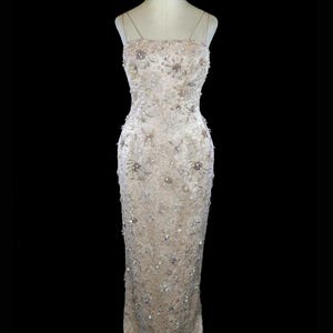 May include: A long, fitted, sleeveless gown with a delicate floral pattern and embellishments. The gown is a light beige color and has a fitted bodice and a flowing skirt.