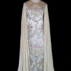 May include: A vintage white gown with floral embroidery and sheer, flowing sleeves. The dress features a high neckline with delicate beading and a long, elegant silhouette. The dress is displayed on a mannequin against a black background.