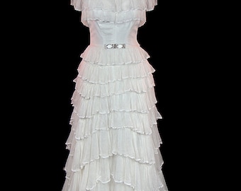 Puede incluir: Un vestido blanco de capas con volantes, con un corpiño ajustado y un escote amplio con volantes. El vestido tiene un cinturón plateado con una hebilla decorativa.