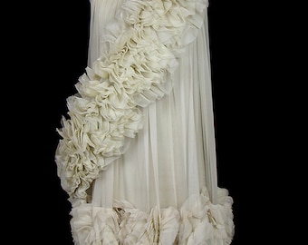 Puede incluir: Un vestido blanco de largo hasta el suelo con un gran detalle de volantes que se envuelve en diagonal sobre el corpiño y continúa por la falda. El vestido está hecho de una tela transparente y tiene un diseño simple y elegante.