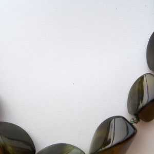 Puede incluir: Un collar hecho de cuentas ovaladas facetadas de color verde oscuro. Las cuentas están enhebradas en un cordón delgado de color verde oscuro.