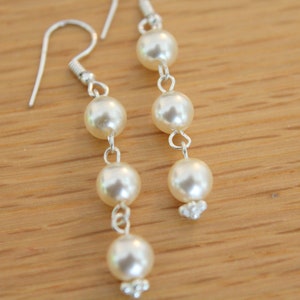 Può includere: Eleganti orecchini pendenti con tre perle rotonde e cremose sospese a ganci color argento. Le perle sono collegate da delicati collegamenti argentati, con un gruppo di piccole perline nella parte inferiore di ogni orecchino. Su una superficie di legno marrone chiaro.