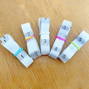Puede incluir: Cinco etiquetas de tela blanca con los números del 1 al 5 impresos en ellas. Cada etiqueta está asegurada con una banda elástica de un color diferente: azul, rosa, amarillo, verde y morado.