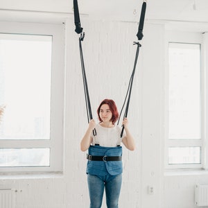 May include: A woman wearing a blue padded harness with a black belt is standing in a white room with a window. She is holding onto two black straps that are attached to the ceiling.