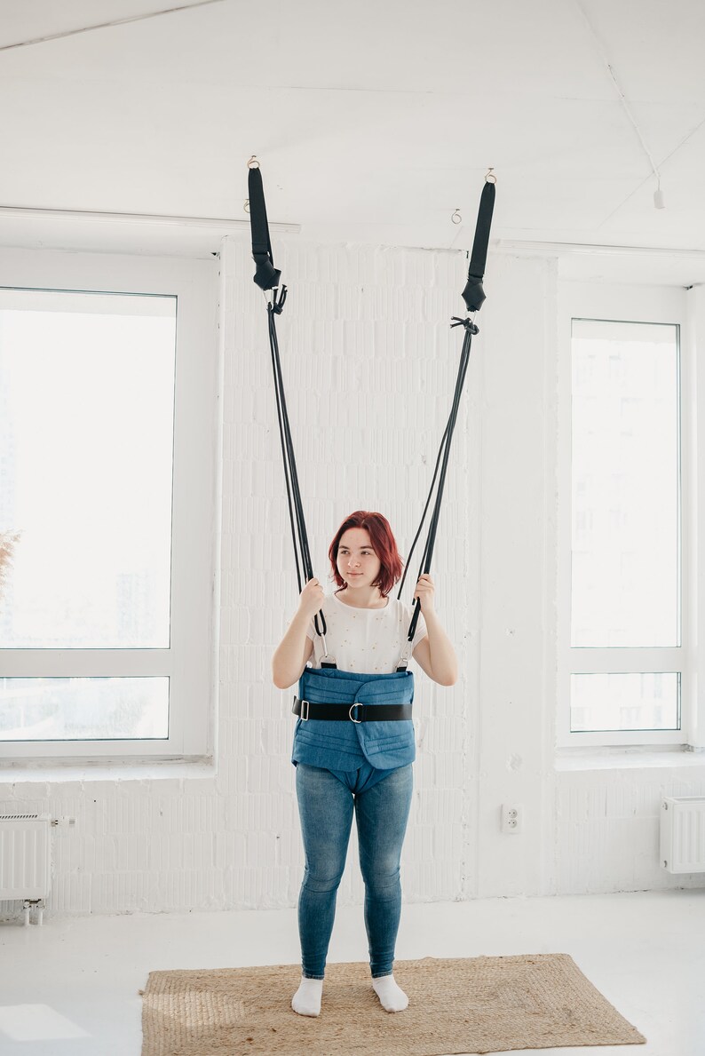 May include: A woman wearing a blue padded harness with a black belt is standing in a white room with a window. She is holding onto two black straps that are attached to the ceiling.