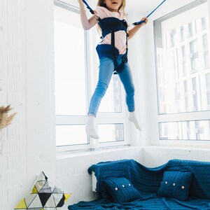 Pode incluir: Uma crian&ccedil;a vestindo uma camiseta rosa claro e cal&ccedil;a jeans azul est&aacute; suspensa no ar por um sistema de arn&ecirc;s azul escuro com cordas el&aacute;sticas azuis presas ao teto. O arn&ecirc;s possui tiras pretas ajust&aacute;veis e fivelas de fixa&ccedil;&atilde;o.