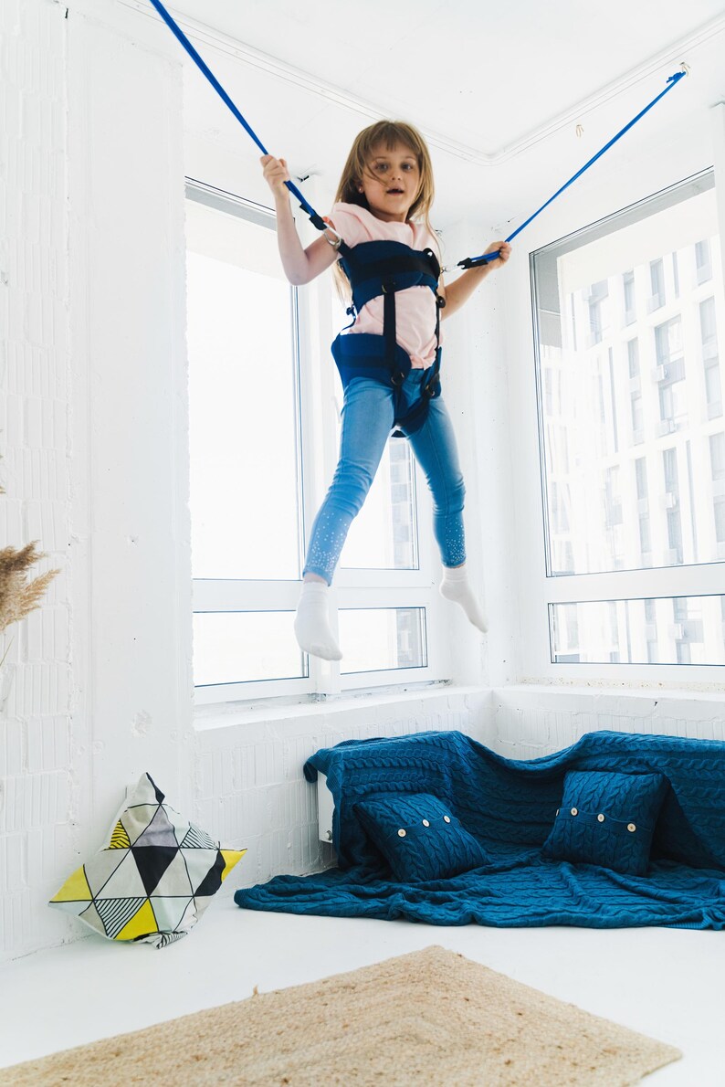 Pode incluir: Uma crian&ccedil;a vestindo uma camiseta rosa claro e cal&ccedil;a jeans azul est&aacute; suspensa no ar por um sistema de arn&ecirc;s azul escuro com cordas el&aacute;sticas azuis presas ao teto. O arn&ecirc;s possui tiras pretas ajust&aacute;veis e fivelas de fixa&ccedil;&atilde;o.