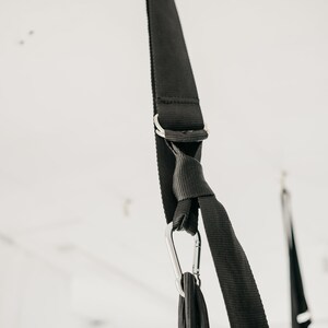 May include: Black straps with a silver carabiner hook hanging from a white ceiling. The straps are tied together in a knot.