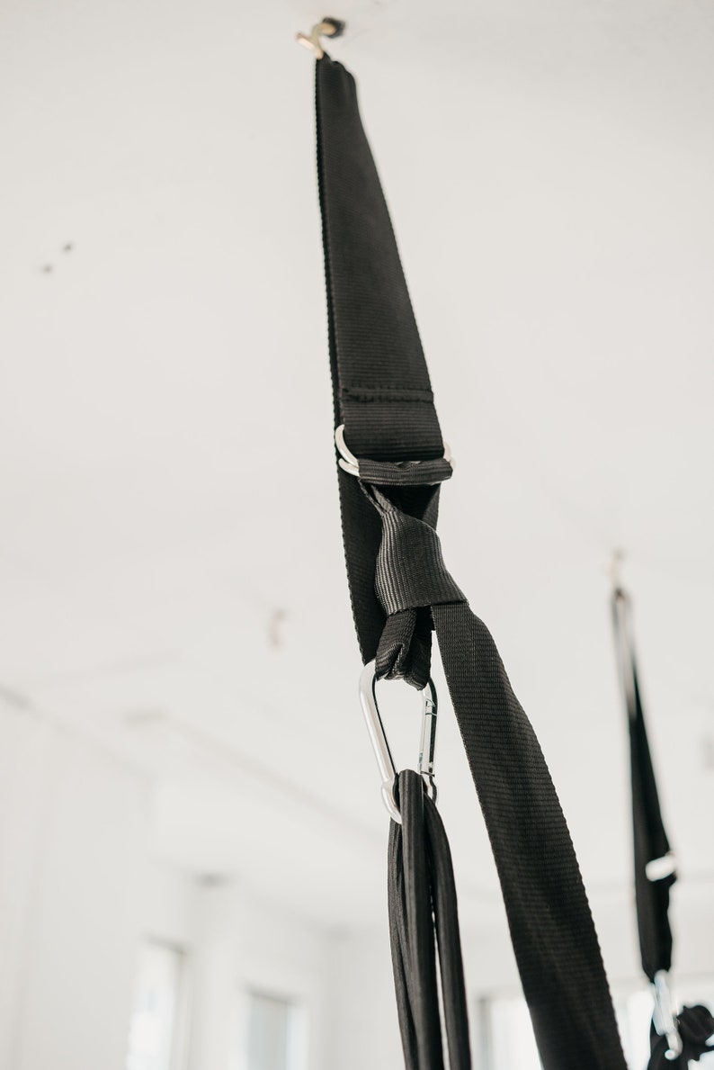 May include: Black straps with a silver carabiner hook hanging from a white ceiling. The straps are tied together in a knot.